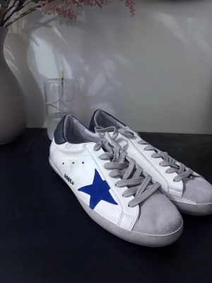 Golden Goose vita sneakers med blå stjärna - Golden Goose sneakers i vittmed grå mockadetaljer på tån, grå snörning och blå stjärna på sidan. Skorna är i väldigt bra skick och kommer med skolådan. Säljes även med original dustbag. Pris kan diskuteras🤝Skriv om du har frågor:)