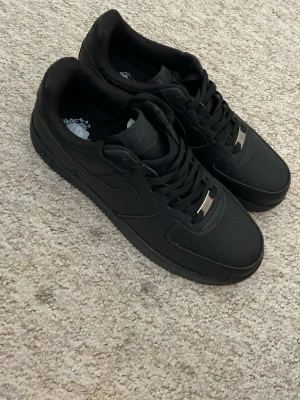 Nike Air Force 1 helsvarta sneakers - Säljer ett par klassiska Nike Air Force 1 i helsvart skinn. Skorna har perforerad tå, svart sula och svarta skosnören med metallbricka framtill. Perfekta för dig som vill ha en clean och tidlös look till vardags. Passar till det mesta och är riktigt bekväma.