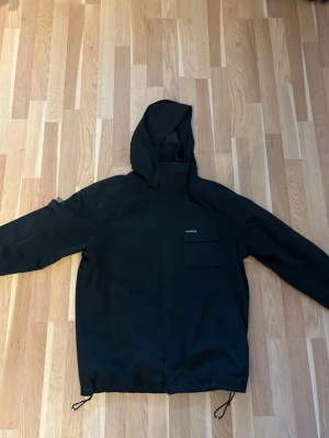 Svart vindjacka från Peak - Säljer en svart vindjacka från Peak med huva och praktisk bröstficka. Jackan har dragsko i nederkant och stängs med dragkedja och vindslå. Perfekt för blåsiga dagar och enkel att matcha med olika outfits.
