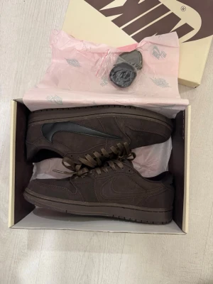 Nike Air Jordan 1 Low Travis Scott - Säljer ett par Nike Air Jordan 1 Low Travis Scott i mörkbrunt mocka med svart swoosh. Skorna har klassisk snörning, perforerad tå och låg profil. Kommer med extra skosnören och originalkartong. Perfekt för dig som vill ha något unikt och stilrent.