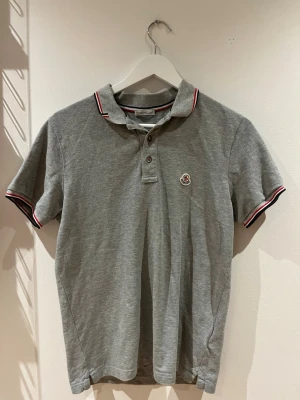 Grå Moncler piké t-shirt S äkta  - Säljer en grå piké t-shirt från Moncler i storlek S. Tröjan har korta ärmar, klassisk krage med röd och vit randig detalj samt Moncler-logga på bröstet. Tillverkad i mjuk bomull och har knappar vid halsen för en clean look.