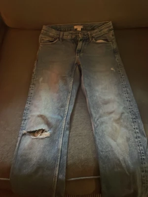 Blå jeans med hål från Gina Tricot! Väldigt snygga med en perfekt midja - Säljer ett par ljusblå jeans från Gina Tricot i storlek 152. Jeansen har ett stort hål på ena knät och klassisk femficksdesign. De är raka i modellen och har en snygg tvättad look.