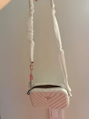 Guess axelväska  - Supersnygg vit axelväska från Guess med quiltad design och guldfärgade detaljer. Väskan har en bred axelrem med tygöverdrag och en kedja med färgglada bokstäver. Dragkedja upptill och logga framtill. Perfekt storlek för det viktigaste! Nypris 1499kr