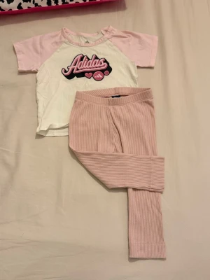 Vit och rosa Adidas t-shirt 12M - Supersöt t-shirt från Adidas i storlek 12 månader. Tröjan är vit med ljusrosa ärmar och har ett stort Adidas-tryck framtill i rosa och svart med små hjärtan och smileys. Perfekt för små trendsetters!