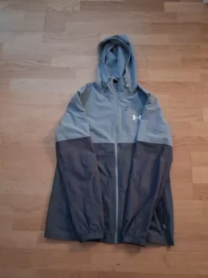 Grå vindjacka från Under Armour - Snygg tvåfärgad grå vindjacka från Under Armour med huva och hel dragkedja. Jackan har en sportig look, sidofickor med dragkedja och logga på bröstet. Perfekt för blåsiga dagar och enkel att slänga på sig över en hoodie.