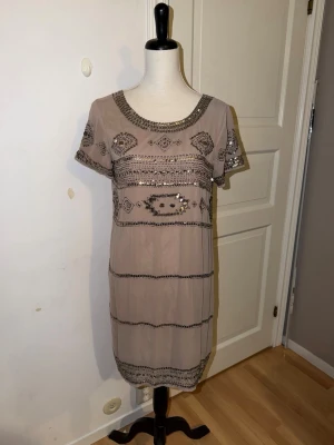 Beige kortklänning med paljetter - Supersnygg beige kortklänning med silverfärgade paljettdetaljer i geometriskt mönster. Klänningen har rund halsringning, korta ärmar och är rak i modellen. Perfekt för dig som vill sticka ut lite extra med glittriga detaljer.