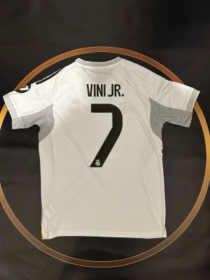 Real Madrid matchtröja Vini Jr. nr 7 - Säljer en officiell Real Madrid matchtröja från Adidas med Vini Jr. och nummer 7 på ryggen. Tröjan är vit med svarta detaljer, gula inslag och klubbmärke på bröstet. Tillverkad i lätt och ventilerande polyester, perfekt för fotbollsplanen.