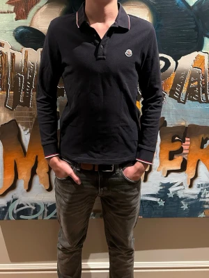 Svart långärmad henley från Moncler - Säljer en svart långärmad henley från Moncler med klassisk krage och knappar framtill. Skjortan har Moncler-logga på bröstet och diskreta rödvita detaljer på krage och ärmslut. Tillverkad i mjuk bomull för skön känsla och snygg passform.