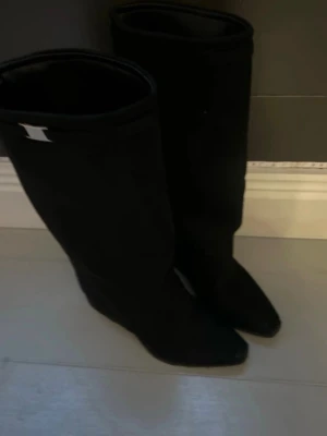 Svarta höga boots i mocka - Säljer ett par svarta höga boots i mjuk mocka med spetsig tå och låg klack. Skorna har en stilren design och en liten metallfärgad detalj upptill på skaftet. Perfekta för utekväll och fester. Säljer dessa för att jag köpte fel storlek💋💕skriv för fler bilder