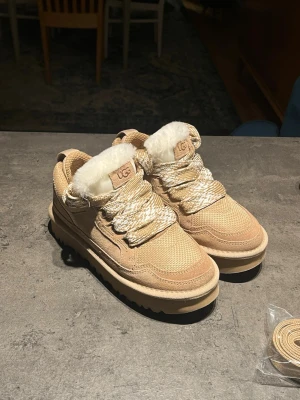 Beige UGG boots med fuskpäls - UGG boots i beige med grov sula och breda snören i beige och vitt. De är endast lite använda. Min flickvän använde dem en kort tid, men de passade tyvärr inte hennes fötter och orsakade skavsår, därför säljs de vidare.  Skorna är i mycket bra skick, rena och väl omhändertagna. Extra skosnören medföljer (se bilder).  Öppet för prisförslag