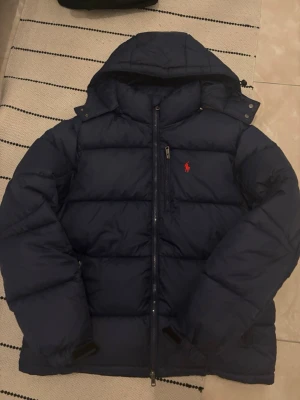 Mörkblå pufferjacka från Polo Ralph Lauren - Säljer en mörkblå pufferjacka från Polo Ralph Lauren med huva och dragkedja framtill. Jackan har det klassiska röda Polo-logotypen broderad på bröstet och är riktigt fluffig och varm. Perfekt för kalla dagar och har en clean, sportig look.