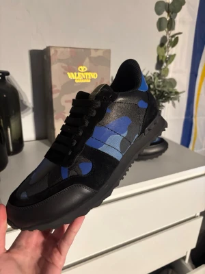 Valentino Garavani Rockrunner sneakers - Säljer ett par Valentino Garavani Rockrunner sneakers i svart och blått med camomönster. Skorna har coola nitar på sulan, snörning och en mix av mocka och skinn. Perfekta för dig som vill sticka ut med en lyxig och edgy look.