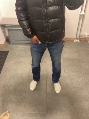 Svart puffer jacka - Säljer en svart pufferjacka med huva och dragkedja framtill. Jackan har två snedställda fickor med dragkedja och en extra ficka på ärmen. Materialet är glansigt och quiltat, perfekt för kalla dagar. Passar dig som vill ha en varm och stilren jacka.