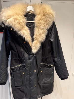 Svart parkas med stor fuskpäls - Säljer en svart parkas från Island med stor fluffig beige fuskpäls runt huvan. Jackan har quiltade detaljer på ärmarna, flera fickor med dragkedjor och knappar framtill. Materialmix av polyester och fuskpäls ger en riktigt snygg look. Pris kan diskuteras!