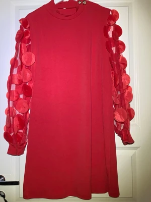 Röd kortklänning med statementärmar - Unik röd kortklänning med transparenta långärmar dekorerade med stora cirkulära applikationer. Klänningen har rund hals med två dekorativa guldknappar på axeln och en rak passform. Perfekt för dig som vill sticka ut med coola detaljer.
