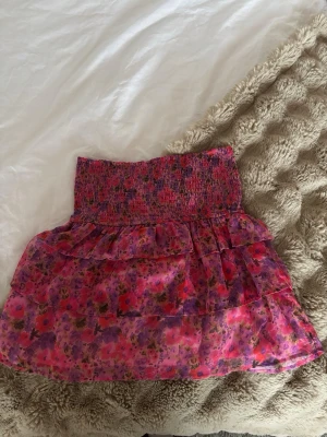 Blommig volangkjol från BikBok - Supersöt kort kjol från BikBok med blommigt mönster i rosa, lila och röda toner. Kjolen har bred smock i midjan och flera lager volanger för en flowy look. Perfekt för sommarens alla äventyr!