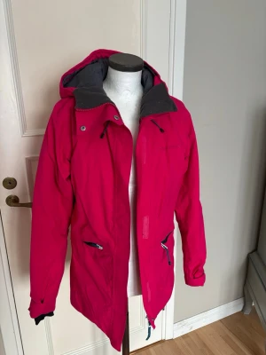 Rosa vindjacka från Didriksons - Snygg rosa vindjacka från Didriksons med huva och grått innerfoder. Jackan har dragkedja framtill, praktiska fickor med dragkedja och justerbara ärmslut. Perfekt för blåsiga dagar och funkar lika bra till jeans som till träningsbyxor.