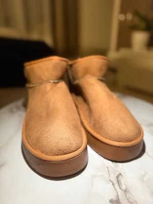 UGG boots i mocka med fårskinn - Säljer ett par klassiska UGG boots i ljusbrun mocka med mjukt och fluffigt fårskinnsfoder. Skorna har rund tå, platt sula och synliga sömmar. Perfekta för kalla dagar när du vill ha både stil och värme.