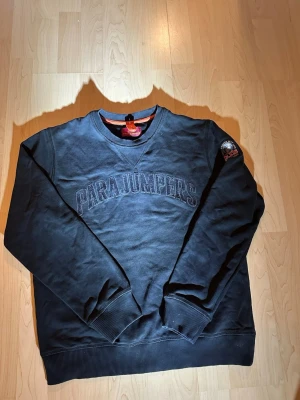 Svart sweatshirt från Parajumpers 13-14 år  - Svart sweatshirt från Parajumpers med broderad logga på bröstet och patch på ärmen. Klassisk rund halsringning, ribbade muddar och nederkant. Tillverkad i mjuk bomull för skön vardagsstil. Snygg detalj med orange band bak.