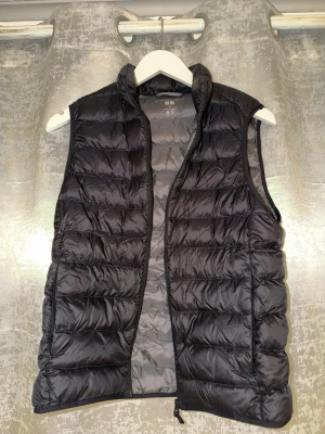 Svart dunväst från Uniqlo - Säljer en svart dunväst från Uniqlo med quiltad design och hög krage. Västen har dragkedja framtill och är lätt och smidig att bära. Perfekt att slänga över en hoodie eller tröja för extra värme.