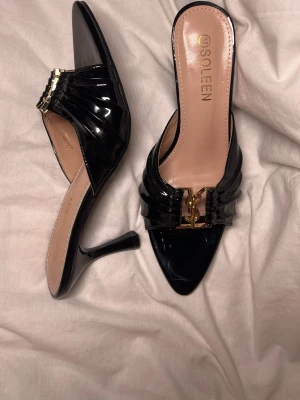 Svarta lackade klackar med gulddetalj - Snygga svarta klackar från Soleen i glansigt lackmaterial med spetsig tå och öppen häl. Skorna har en elegant guldfärgad detalj framtill och smal hög klack. Jättebekväma. Har i storlek 36 samt 37, och även beiga. Endast provade, storlek 37 har dock en liten repa som syns på bild men inget som märks av. Blev lite för små för mig ☹️