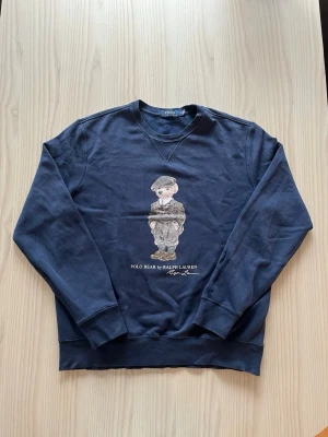 Mörkblå sweatshirt Polo Ralph Lauren - Säljer en mörkblå sweatshirt från Polo Ralph Lauren med den ikoniska Polo Bear-figuren tryckt på bröstet. Tröjan har rund halsringning och långa ärmar. Tillverkad i mjuk bomull för skön känsla och stilren look. 