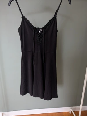 Svart kort klänning med spetsdetaljer - Supersöt svart kort klänning med tunna axelband och v-ringning. Klänningen har spetsdetaljer längs urringningen och snörning framtill för extra stilpoäng. Materialet känns mjukt och stretchigt, perfekt för dig som gillar en enkel men ändå romantisk look.