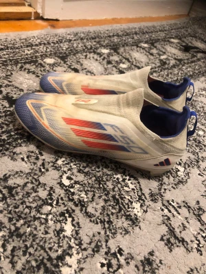 Adidas F50 Pro Laceless LL Euro Edition  - Säljer ett par Adidas F50 fotbollsskor i vitt med blå och röda detaljer. Skorna har en strömlinjeformad design utan snörning, med mönstrad ovandel och dobbar i rött och vitt. Perfekta för dig som vill sticka ut på planen.