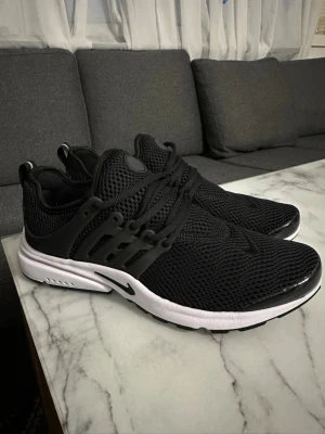 Svarta Nike Presto sneakers - Säljer ett par svarta Nike Presto sneakers med vit sula och svart mesh-överdel. Skorna har snörning, rund tå och en sportig vibe. Sulan är i syntetmaterial och ovandelen är ventilerande mesh för extra komfort. Perfekta för dig som gillar stilrena och bekväma sneakers.