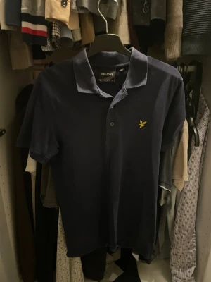 Mörkblå piké från Lyle & Scott - Säljer en mörkblå pikétröja från Lyle & Scott med klassisk krage och två knappar framtill. Tröjan har korta ärmar och den ikoniska gula logon broderad på bröstet. Perfekt till jeans eller shorts för en clean look.