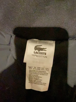 Svart hoodiejacka från Lacoste - Säljer en svart hoodiejacka från Lacoste. Jackan har dragkedja framtill, huva och det klassiska Lacoste-märket på bröstet. Perfekt för dig som vill ha en stilren och sportig look med en bekväm passform.