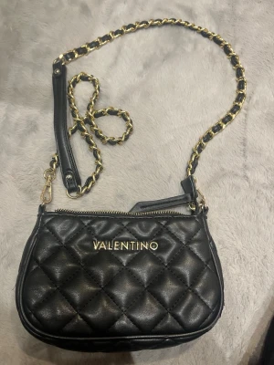Valentino väska - Valentino med guldfärgade detaljer och kedjerem. Väskan är i skinnimitation och har dragkedja upptill samt flera fack inuti för kort och småsaker. Perfekt storlek för det viktigaste och riktigt snygg design.