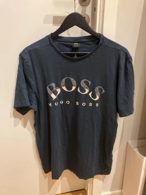 Mörkblå Hugo Boss t-shirt i bomull - Snygg mörkblå t-shirt från Hugo Boss med stort vitt BOSS-tryck på bröstet. Klassisk passform och rund halsringning. Tillverkad i mjuk bomull för skön känsla och stilren look.