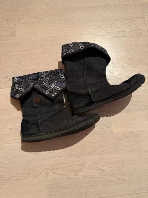 Mörkblå UGG boots med mönstrat foder - Säljer ett par mörkblå (jeanslook) UGG boots i textil med vikbar skaftkant som har ett coolt mönster i vitt, grått och blått. Sulan är platt och robust, och bootsen har dekorativa knappar på sidan. Perfekta för dig som vill ha både stil och komfort.