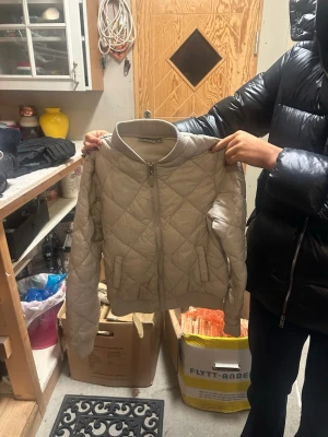 Beige quiltad bomberjacka Rosetti - Säljer en beige quiltad bomberjacka från Rosetti med ribbade muddar och dragkedja framtill. Jackan har ett rutigt mönster i quiltningen och två fickor fram. Perfekt för dig som vill ha en clean och stilren look.