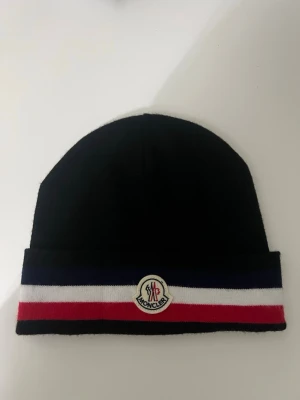 Moncler Mössa -  Moncler mössa i klassisk modell. Svart med ikoniska ränder och Moncler-logga framtill. Använd men i fint skick, inga hål eller fläckar. Stretchig och bekväm passform, passar både dam och herr (unisex). Perfekt för höst och vinter.