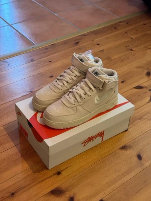 Nike Air Force 1 Mid Fossil x Stüssy - Nike Air Force 1 Mid SP i samarbete med Stüssy i färgen Fossil. Skorna har en beige canvas-överdel, klassisk snörning, kardborreband runt ankeln och vit swoosh. Snygg och clean siluett med rund tå och platt sula. Perfekt för dig som gillar streetwear och unika collabs.