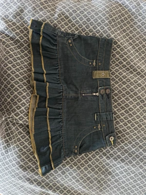 Mörkblå volangkjol från MNG Jeans - Supersnygg mörkblå jeanskjol från MNG Jeans med volangkant och coola detaljer som fickor, knappar och dragkedja framtill. Kjolen har kontrastsömmar i gult och markerad midja. Perfekt för dig som vill ha en edgy och unik look.