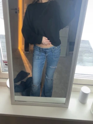 Lågmidjade blå bootcut jeans - Säljer ett par ljusblå jeans med låg midja och bootcut-ben. Jeansen har slitna detaljer och hål vid knäna för en trendig look. Klassisk femficksmodell och tillverkade i jeansmaterial. Perfekta för dig som gillar en avslappnad och cool stil.