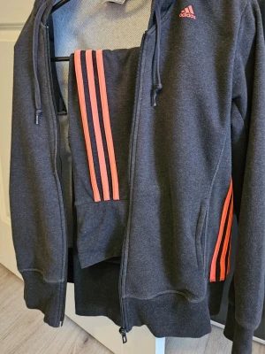 Mörkgrå Adidas träningsväst med rosa ränder - Säljer en mörkgrå träningsväst från Adidas med huva och dragkedja. Västen har de klassiska tre rosa ränderna längs sidorna och en rosa Adidas-logga på bröstet. Tillverkad i mjuk bomullsblandning, perfekt för sportiga outfits.