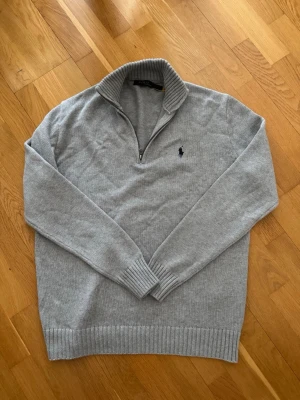 Ljusgrå stickad tröja Polo Ralph Lauren - Ljusgrå stickad tröja från Polo Ralph Lauren med halv dragkedja och hög krage. Tröjan har ribbade muddar vid ärmslut och nederkant samt den klassiska mörkblå logotypen broderad på bröstet. Perfekt för lager på lager och stilren look.
