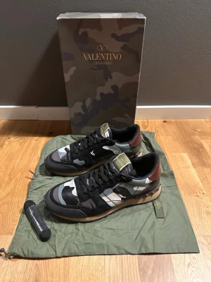 Valentino Rockrunner camo sneakers - Snygga Valentino Rockrunner sneakers med camouflage-mönster i svart, grått, vitt och olivgrönt. Skorna har svarta snören, rund tå och detaljer i mocka och skinn. Klassisk Valentino Garavani-logga på plösen och cool kontrast på sulan.