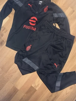 AC Milan träningsset från Puma - Snyggt svart träningsset från Puma med AC Milan-logga och röda detaljer. Setet består av långärmad tröja med halv dragkedja och matchande byxor, båda med grå reflexdetaljer. Perfekt för fotbollsträning eller sportiga dagar. Pris för nytt är ca 900-1000kr.