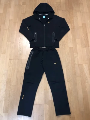 Nocta tech fleece tracksuit - Svart Nike NOCTA set bestående av zip-hoodie med huva och matchande byxor. Stilren och exklusiv design med guldfärgade Nike-loggor och NOCTA-detaljer. Tech-material med dragkedjefickor. ✔ Original Nike NOCTA ✔ Färg: Svart med gula detaljer ✔ Skick: nästan nytt – endast använd 2–3 gånger ✔ Säljs som set (tröja + byxor) Passar perfekt för streetwear, vardag eller träning. 🔥 Skicka gärna meddelande vid frågor eller om du vill ha fler bilder.