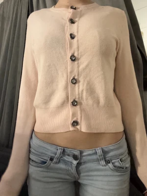 Ljusrosa kofta med blommiga knappar - Söt ljusrosa kofta med långa ärmar och blommiga dekorativa knappar framtill. Tröjan har en croppad passform och ribbade muddar vid ärmslut och nederkant. Perfekt för att addera en feminin touch till din outfit.