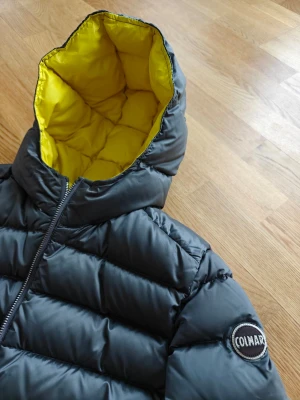 Mörkgrå Colmar dunjacka med huva - Snygg mörkgrå dunjacka från Colmar med gul insida på huvan. Jackan har quiltad design, dragkedja framtill och två sidofickor. Perfekt för kalla dagar och ger en sportig look. Superfin,varm jacka. Ren, nytvättad inför försäljningen. Ett litet märke finns ,en centimeter brett märke där kedjan fastnat, så det blev tråd dragning. Inget som syns vid första anblicken, eller stör funktion. För övrigt väldigt fint skick. Storlek 42, italiensk storlek,men upplevs som 34 / xs.