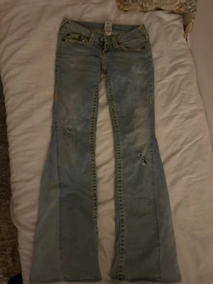 Ljusblå true religion bootcut jeans  - Säljer ett par ljusblå jeans från True Religion med tydliga gula kontrastsömmar och klassiska bakfickor med broderad logga. Modellen har låg midja och bootcut-ben. Jeansen har slitningar och detaljerade nitar vid fickorna för en extra cool look. Storlek 26 men passar mig hyfsat som har 28/29