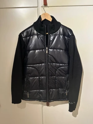 Svart cardiganjacka med stickade ärmar - Snygg svart cardiganjacka med glansig, quiltad framsida och stickade ärmar samt rygg. Jackan har hög krage och dragkedja framtill, samt två fickor med dragkedja. Perfekt för dig som vill ha en modern och mysig look. Helt ny.