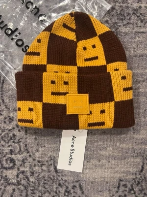 Acne Studios beanie   - Stickad mössa från Acne Studios i brunt och gult med rutigt mönster och smiley-ansikten. Mössan har uppvikt kant och en broderad patch framtill med Acne Studios klassiska ansikte. Perfekt för dig som vill sticka ut med en cool accessoar.