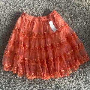 Orange volangkjol med spetsdetaljer - En fluffig orange kjol med flera lager volanger och spetsdetaljer. Kjolen har resår i midjan och är tillverkad i ett lätt, transparent material som ger en luftig känsla.